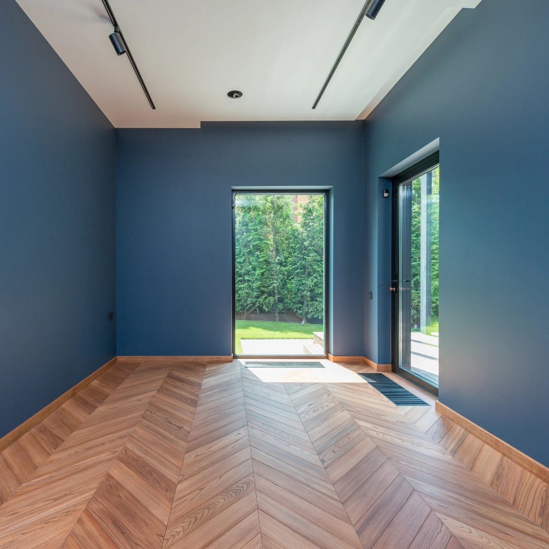 Raum mit dunkelblauen Wänden, weißer Decke, Holzboden im Chevron-Muster und bodentiefen Fenstern zum Garten.