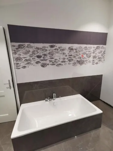 Weiße Badewanne mit dunkler Fliesenverkleidung vor einer Wand mit dekorativem Fischmuster