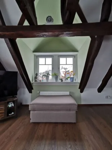 Dachgeschossraum mit dunklen sichtbaren Holzbalken, hellgrüner Fensternische und weißen Dachschrägen
