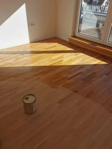 Parkettboden während der Oberflächenbehandlung mit unterschiedlich glänzenden Bereichen und einer Dose auf dem Boden